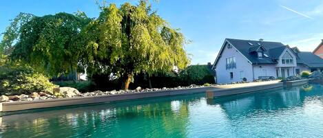 Una piscina con muelle de madera, una casa blanca y un árbol grande.