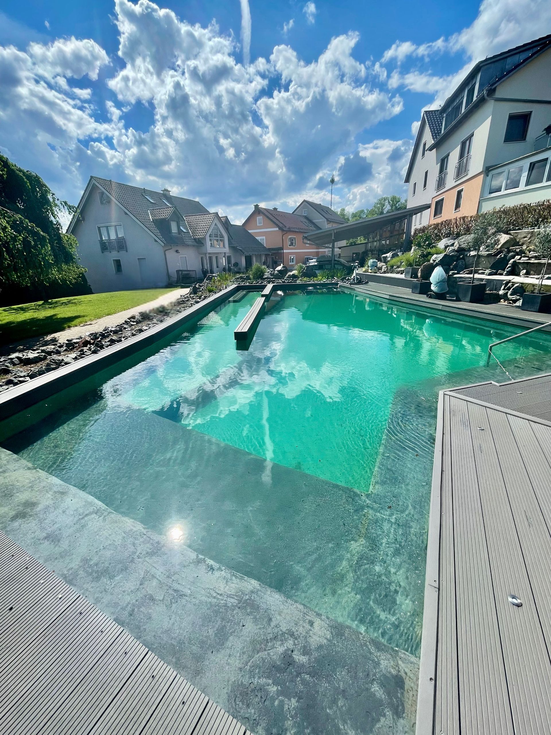 Ein Schwimmbecken mit klarem Wasser, umgeben von einer Holzterrasse, neben Wohngebäuden.