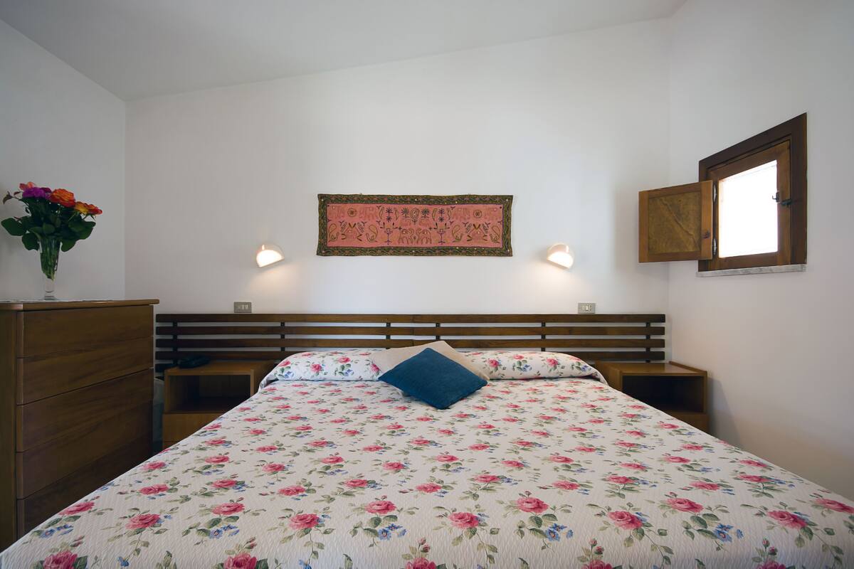 A bedroom with a floral bedspread, a wooden headboard, a bedside table, a nightstand, a wall-mounted lamp, and a window with a wooden frame.