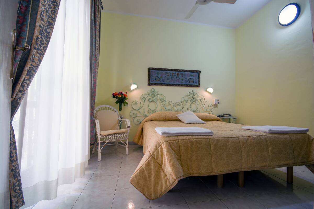 A hotel room with a bed, a chair, a nightstand, and a wall-mounted light fixture.