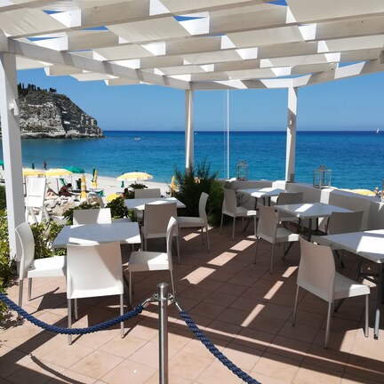 A covered outdoor seating area with white tables and chairs, overlooking a beach with umbrellas and people.