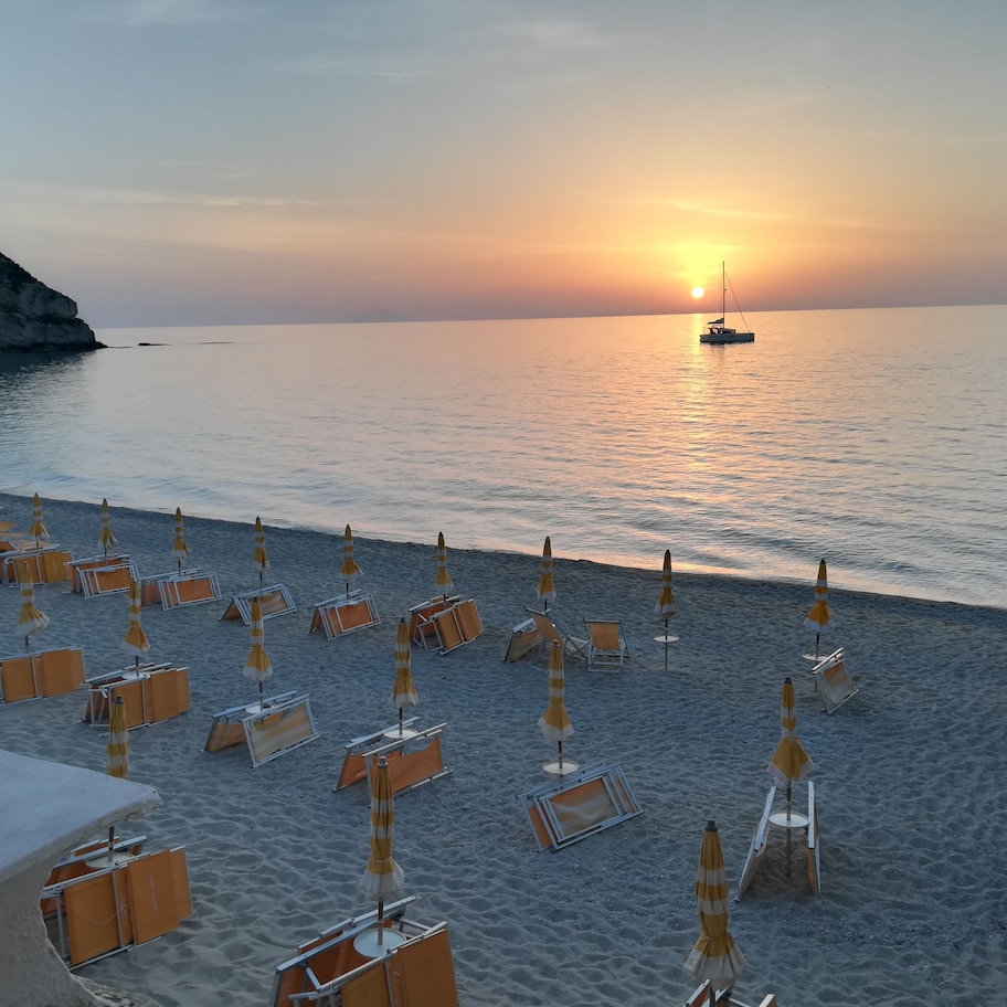 A beach with rows of sun loungers and umbrellas, a boat in the water, and a sunset in the background.
