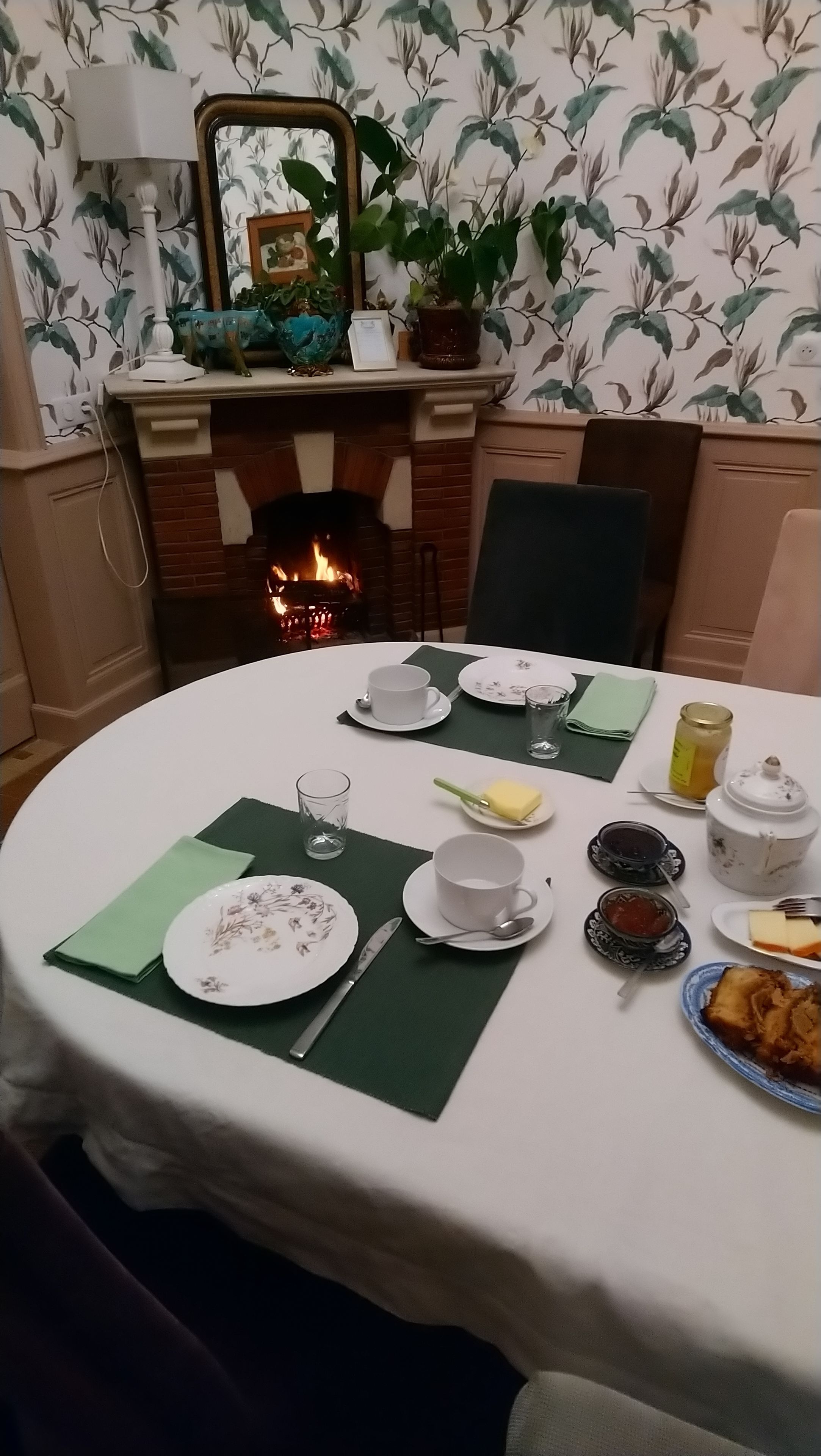 Une table à manger est dressée pour quatre personnes, avec des assiettes, des verres et des couverts. On aperçoit une cheminée au fond.