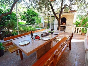 An outdoor dining area with a stone table and wooden benches, surrounded by greenery and a stone fireplace.