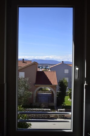 Une vue prise de l’intérieur d’un bâtiment, à travers une fenêtre, donnant sur un quartier résidentiel avec des maisons, un mur de pierre et un ciel bleu dégagé.