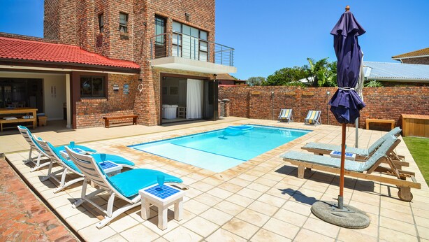 A swimming pool with lounge chairs and a covered area, surrounded by a brick building and a grassy lawn.
