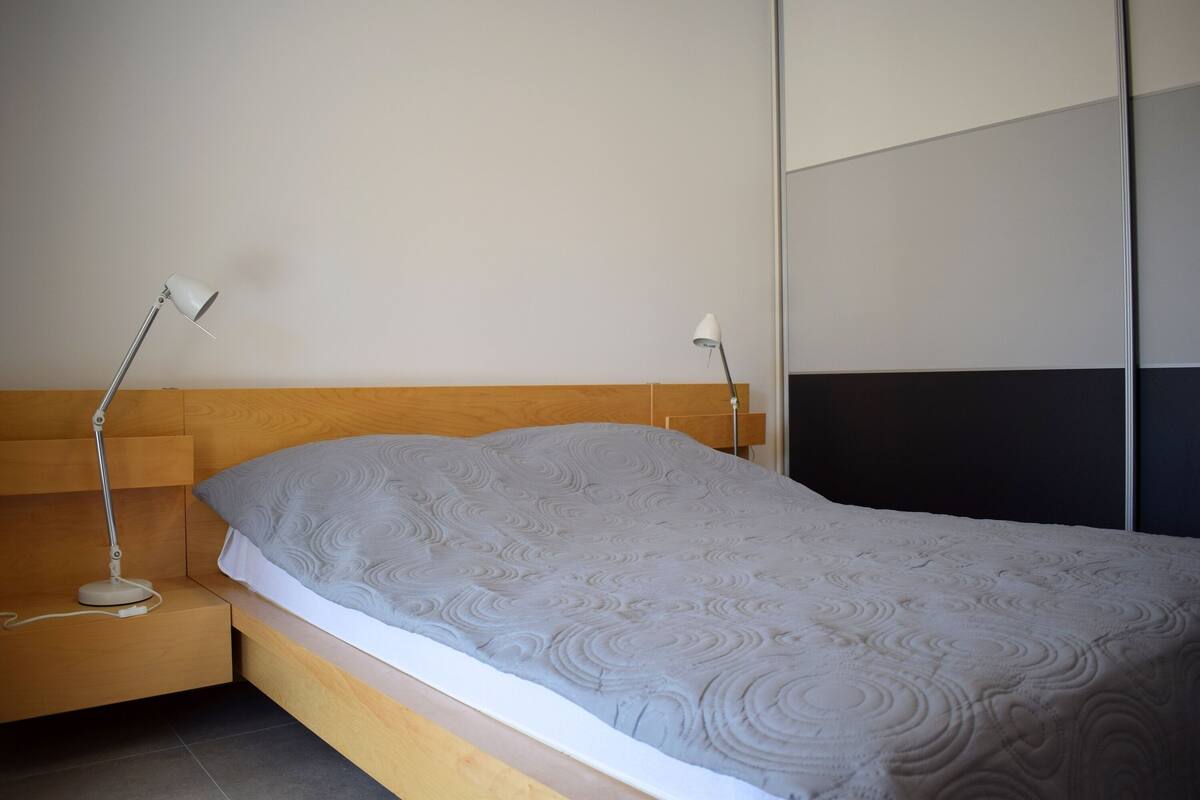 A modern bedroom with a wooden headboard, a grey patterned bedspread, and two bedside lamps.