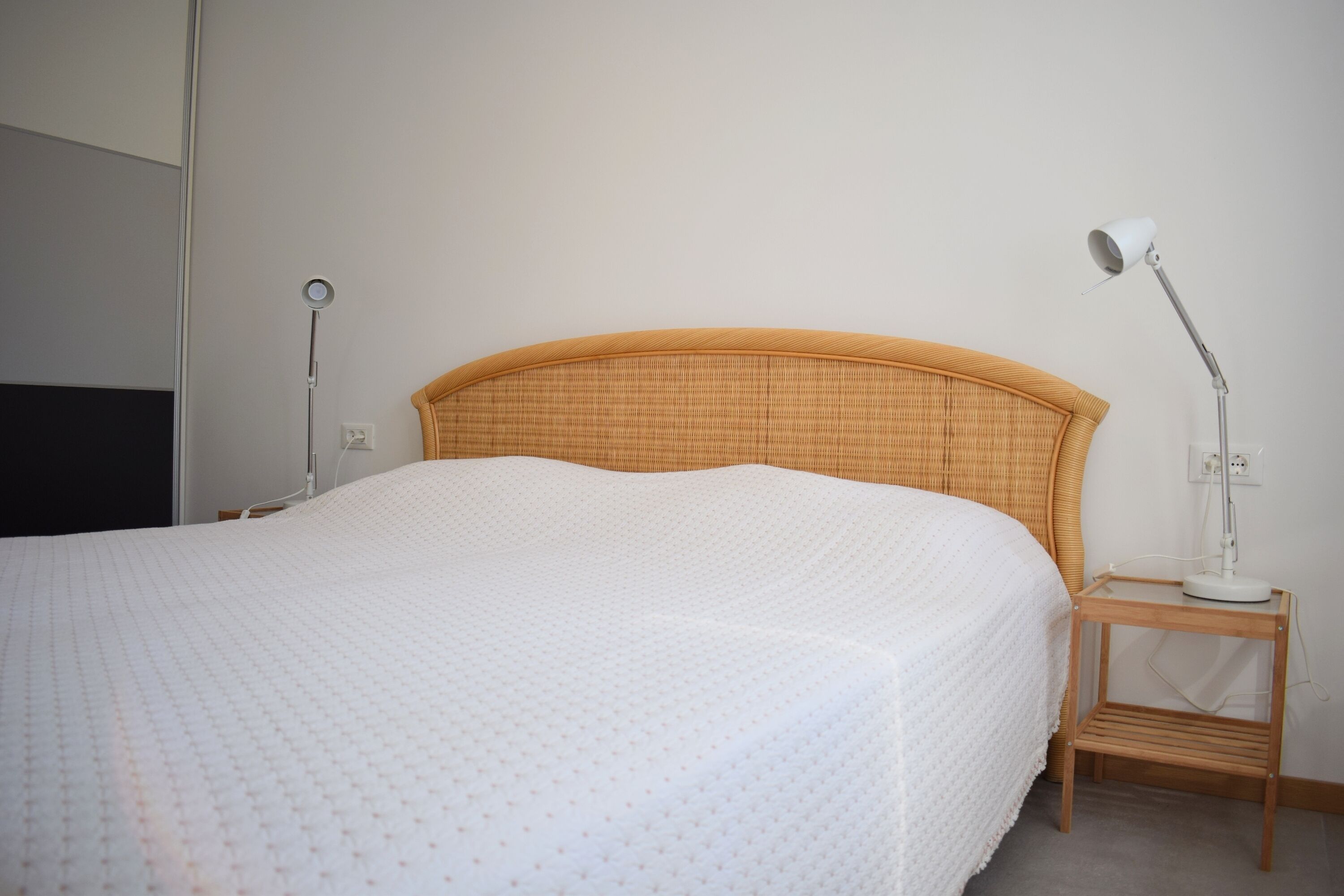 Una cama bien hecha con una colcha blanca texturizada, un cabecero de madera y una mesita de noche con una lámpara.