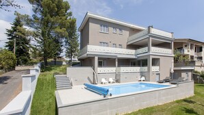 A modern two-story building with a swimming pool, outdoor seating, and a stone wall.