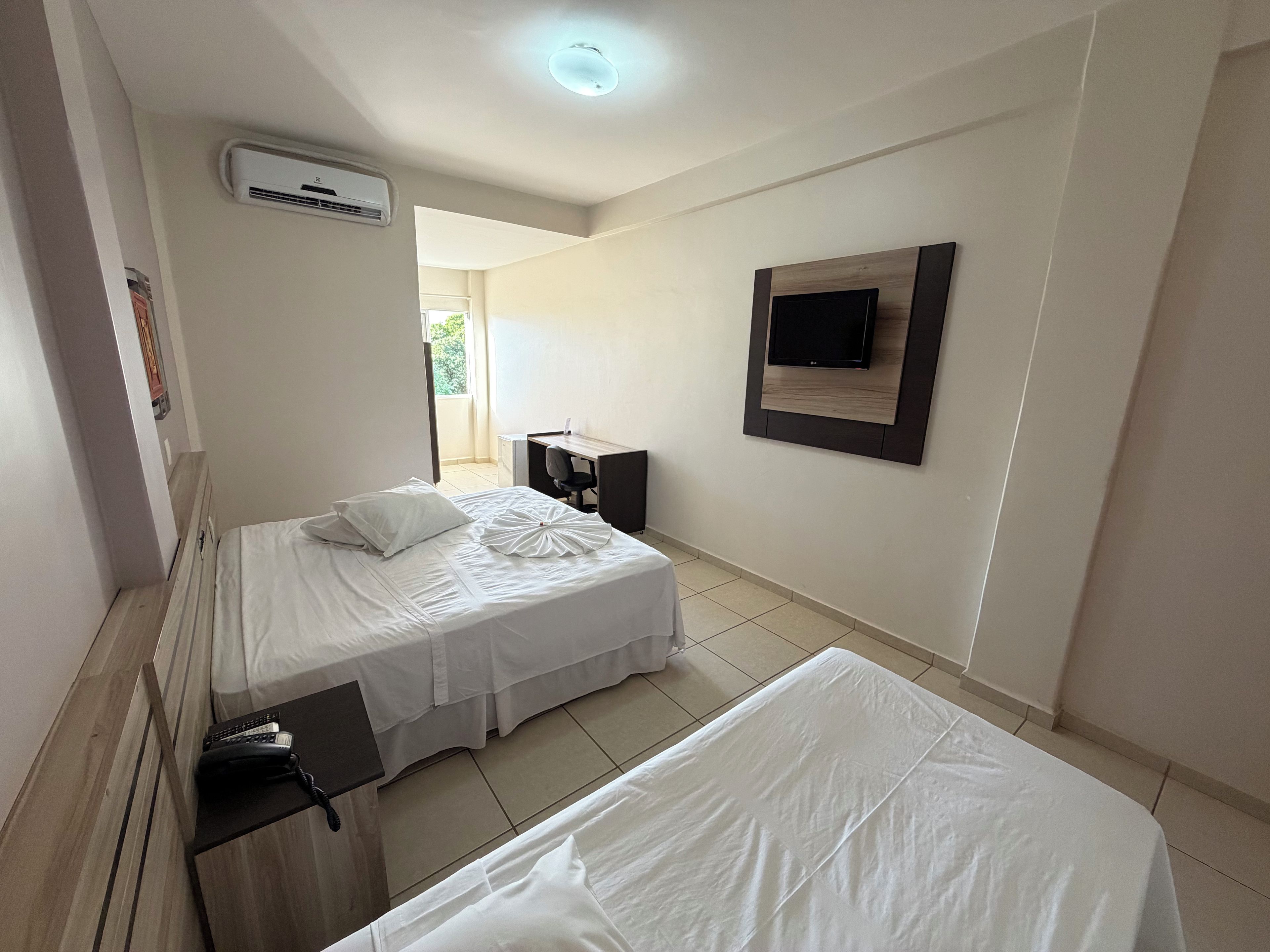 A hotel room with two beds, a desk, a chair, a TV mounted on the wall, and an air conditioning unit.