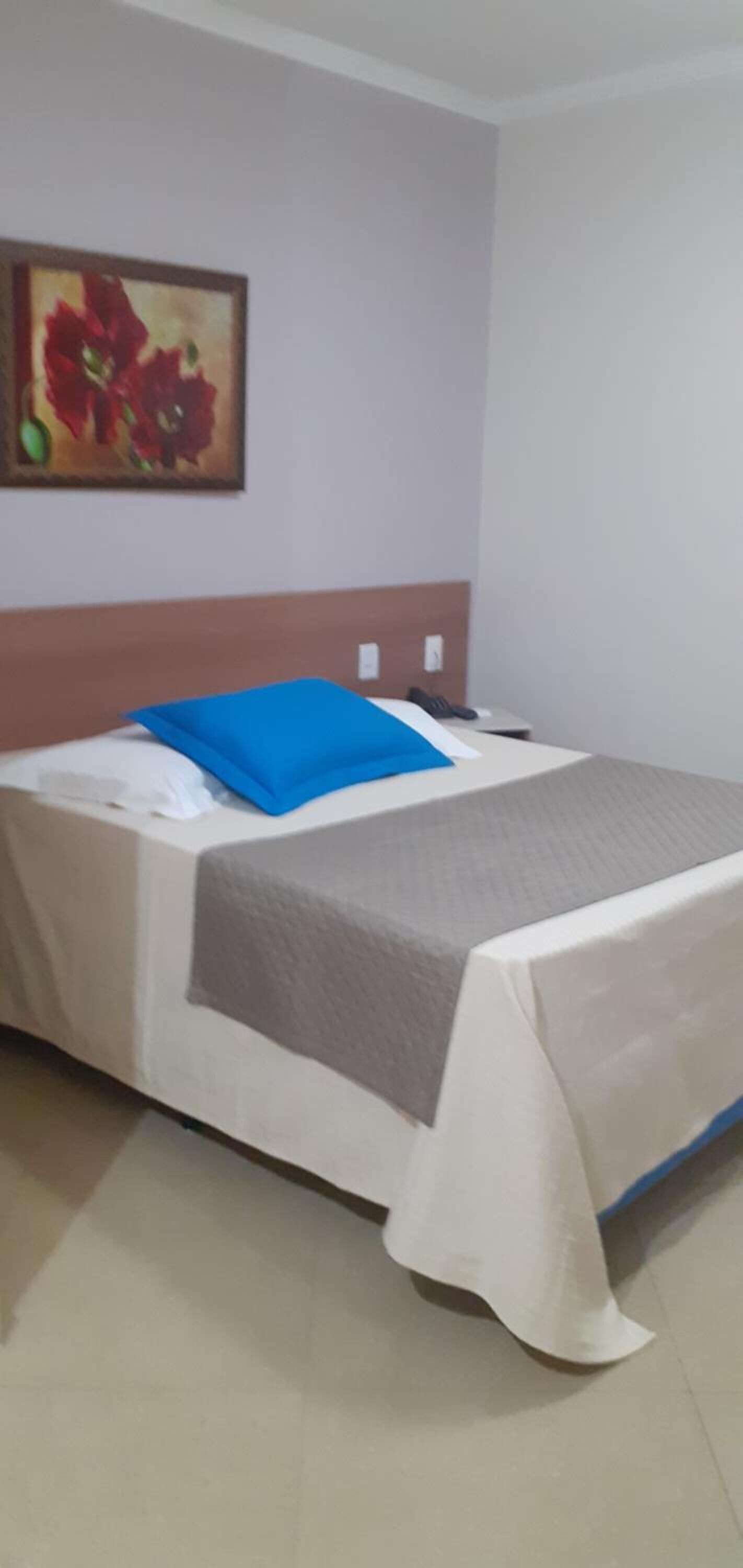 A neatly made bed with a blue pillow and a grey blanket, a painting on the wall, and a white headboard.