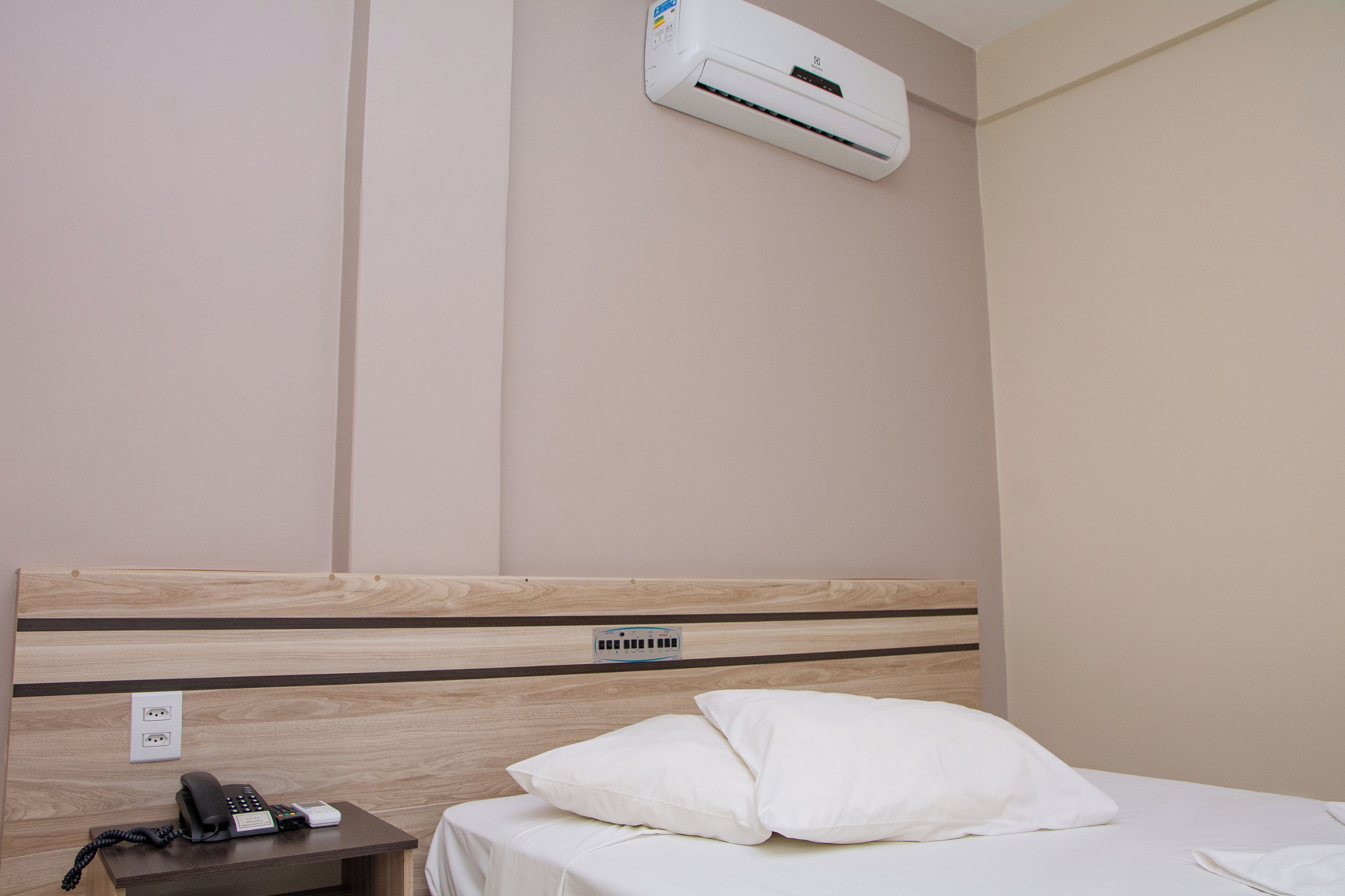 A hotel room with a wooden headboard, a white bed, a white pillow, a bedside table with a phone and a remote control, and an air conditioning unit mounted on the wall.