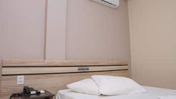 A hotel room with a wooden headboard, a white bed, a white pillow, a bedside table with a phone and a remote control, and an air conditioning unit mounted on the wall.