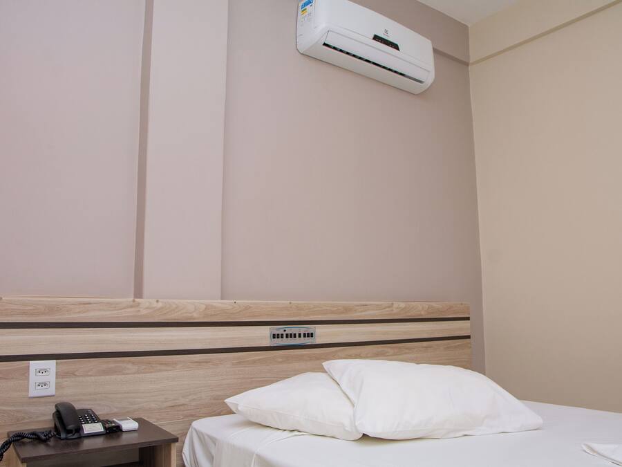 A hotel room with a wooden headboard, a white bed, a white pillow, a bedside table with a phone and a remote control, and an air conditioning unit mounted on the wall.