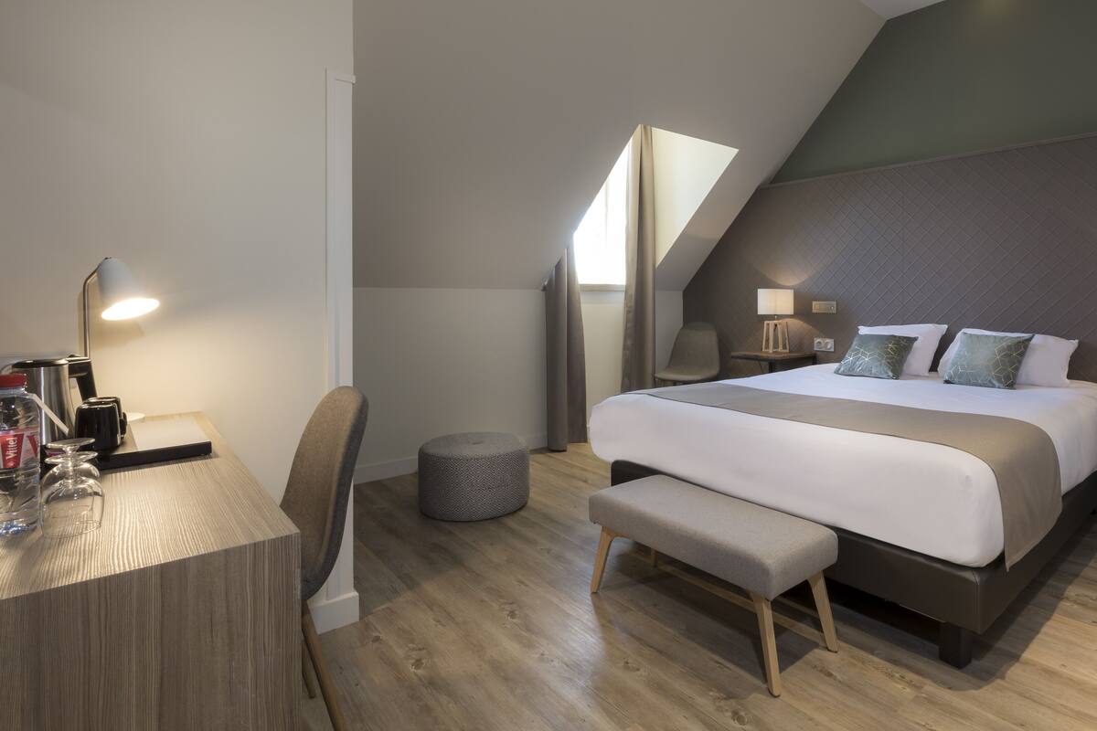 Un dormitorio moderno con una cama grande, un escritorio con lámpara, una silla y un otoman redondos.