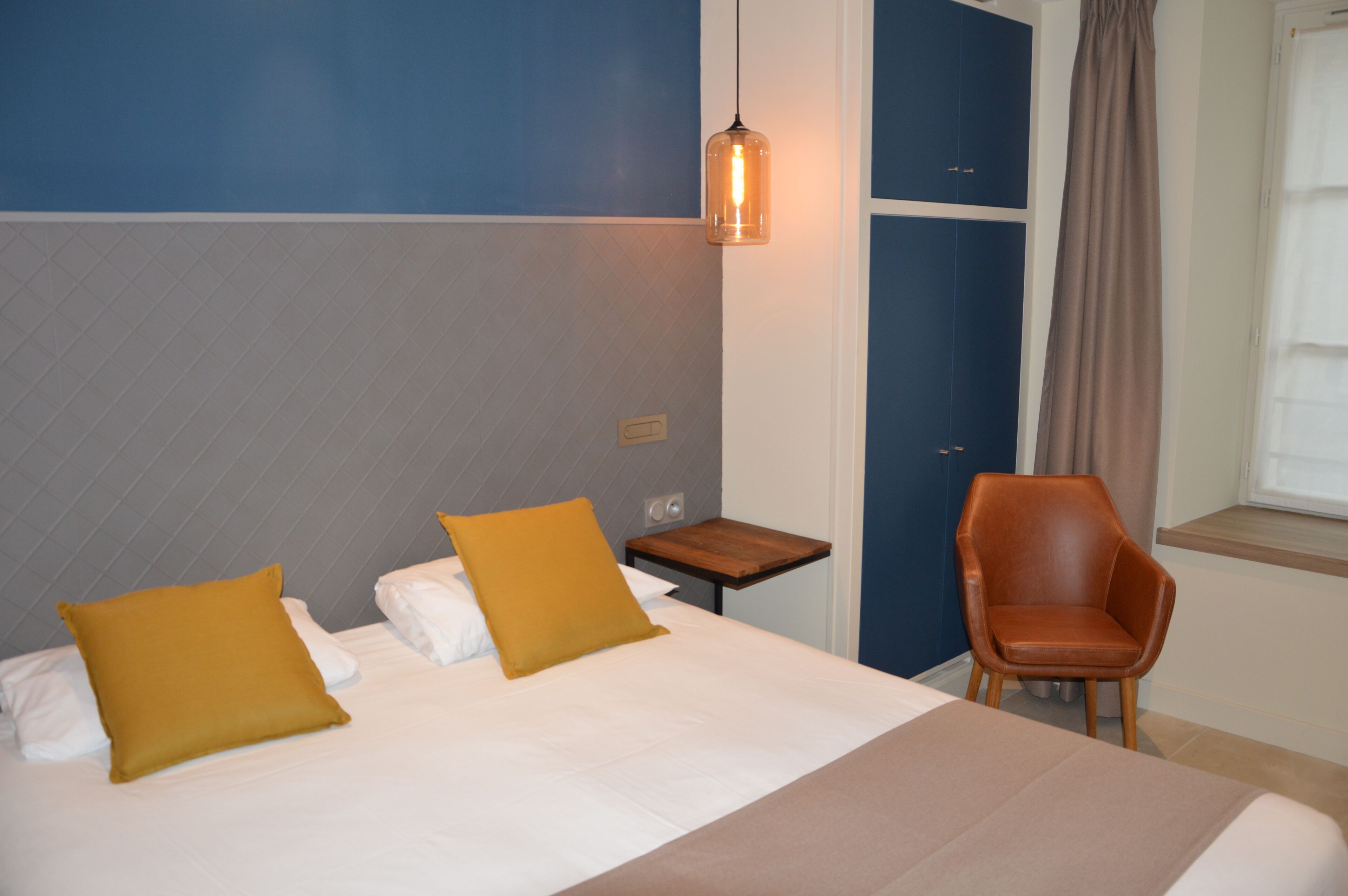 A hotel room with a bed, two pillows, a nightstand, a chair, a hanging light, and a window with curtains.