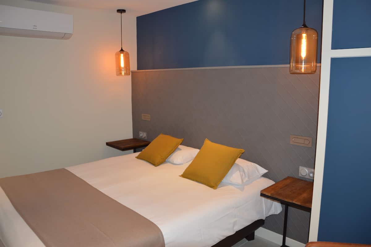 Habitación de hotel con cama, dos mesitas de noche, aire acondicionado instalado en la pared y dos lámparas colgantes.