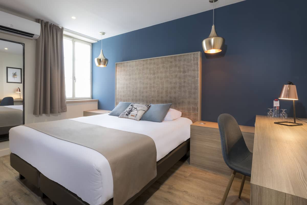 Habitación de hotel moderna con una cama grande, un escritorio, una silla y un amplio ventanal con cortinas.