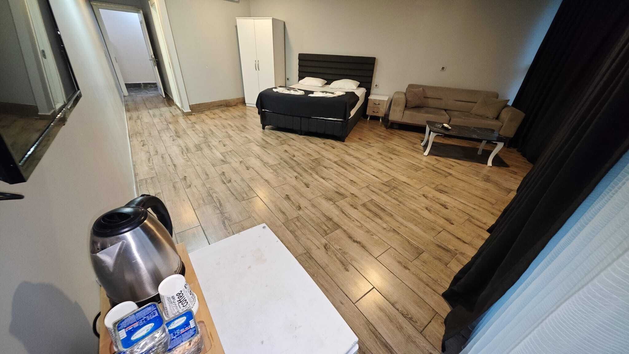 Habitació d'hotel amb sòl de fusta, un llit amb capçalera negra, un sofà beix, una taula petita i un armari blanc.