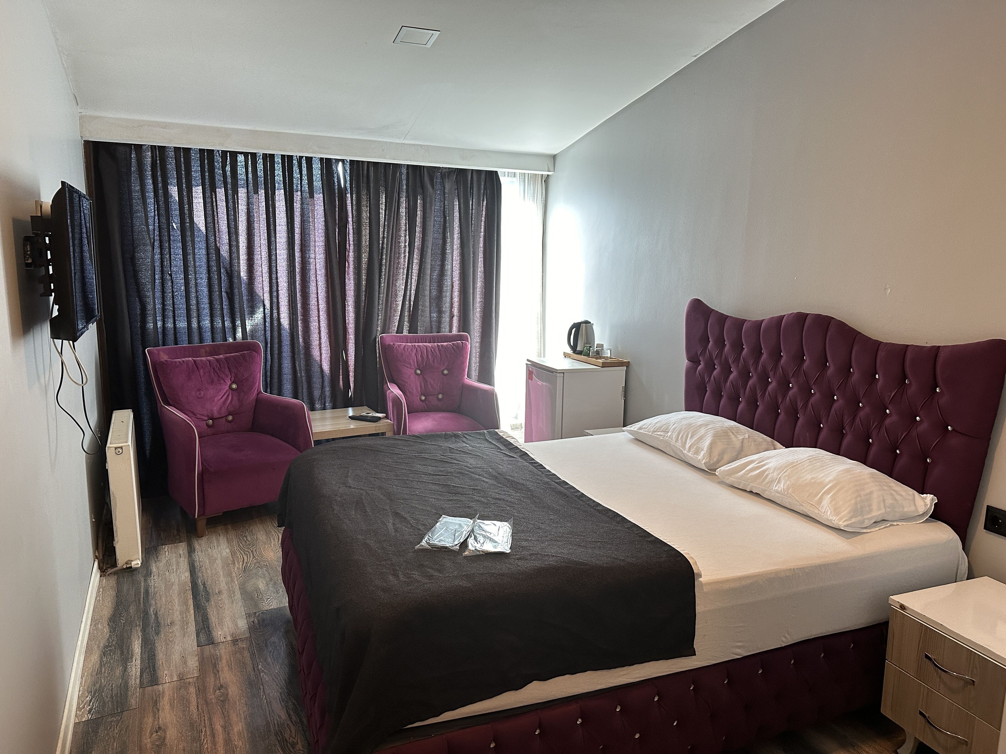 Habitación de hotel con cama, dos sillas, una mesita, televisor en la pared y ventana con cortinas.