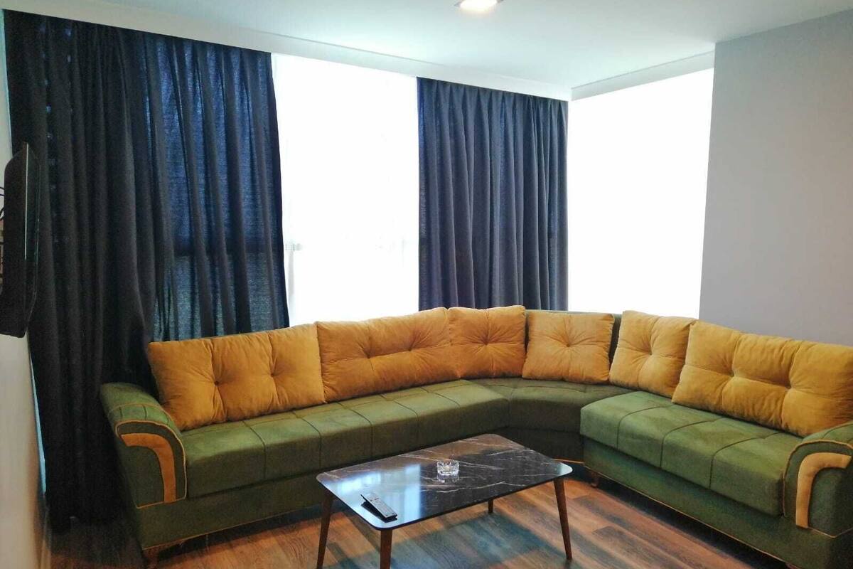 A living room with a green sectional sofa, a glass coffee table, and wooden flooring.