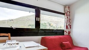 Una habitación con una mesa de madera preparada para dos personas, un sofá rojo y una ventana que ofrece vistas a un paisaje montañoso.