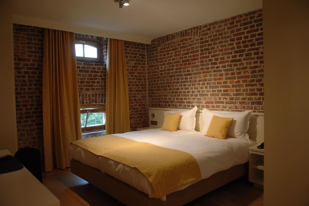 Habitación de hotel con pared de ladrillo, una cama grande con ropa de cama blanca y amarilla, una mesita de noche y una ventana con cortinas.