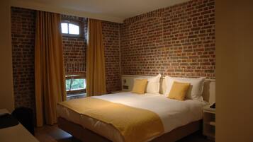 A hotel room with a brick wall, a large bed with white and yellow bedding, a nightstand, and a window with curtains.