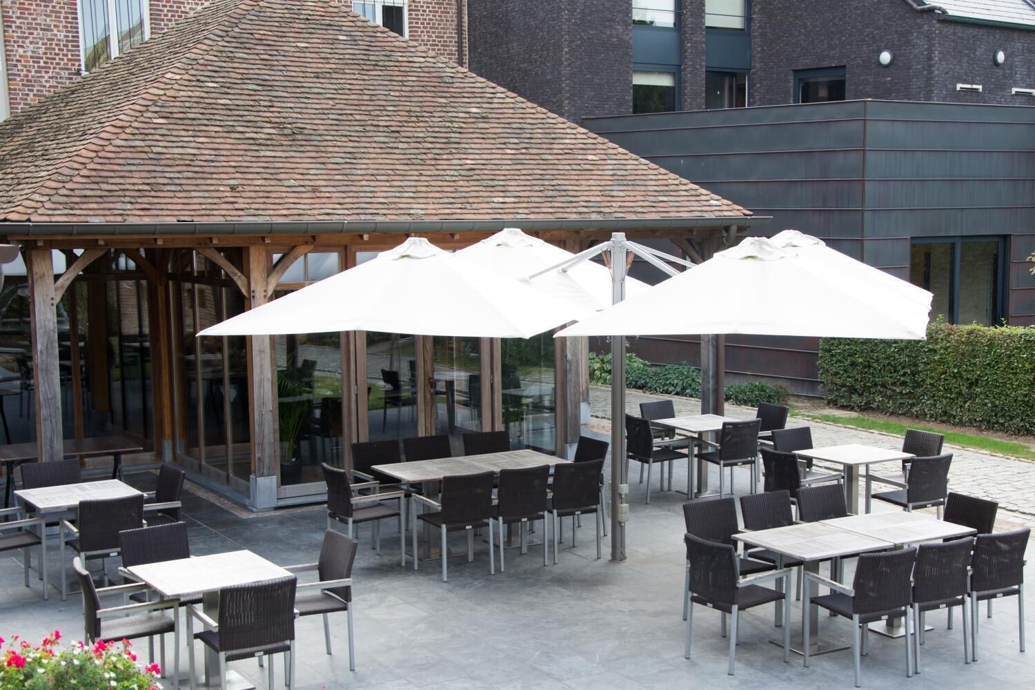 Een terras buiten met tafels en stoelen, afgedekt door parasols, en een glazen constructie met een bakstenen gebouw op de achtergrond.