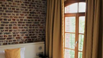 Una habitación de hotel con pared de ladrillo, un ventanal con cortinas, una cama con ropa blanca y una mesita de noche con lámpara.