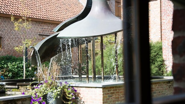 A water feature with a curved metal structure and a fountain in front of a brick building. - Hostellerie de Biek (Aalst)