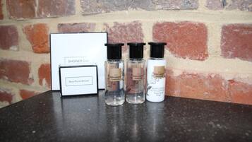 Un gorro de ducha, una esponja para lustrar zapatos y tres botellas de productos The Perfumer's Garden sobre una mesa frente a un muro de ladrillo.