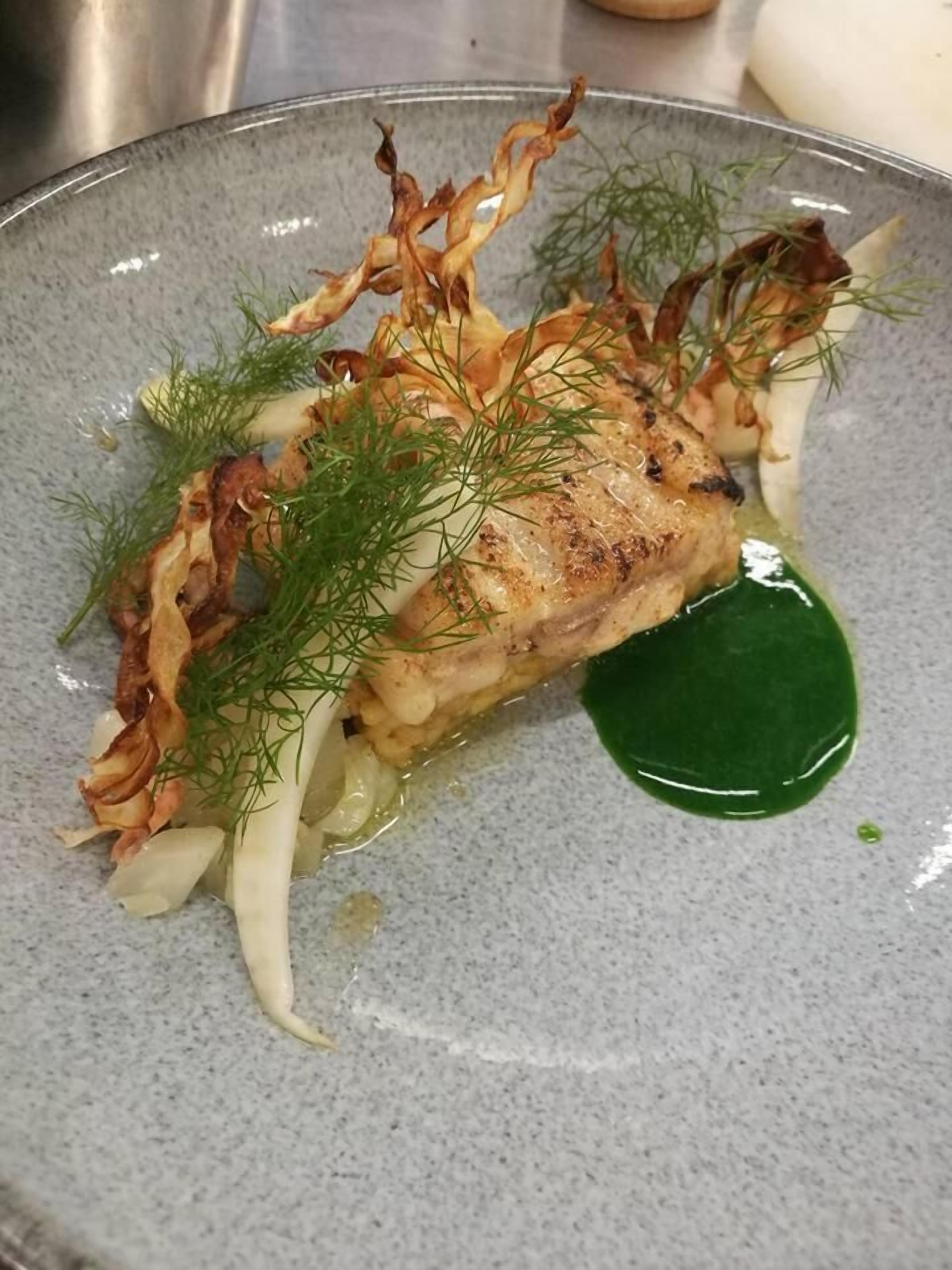 A plate of food with a piece of grilled fish, garnished with dill and crispy elements, served on a speckled ceramic plate.