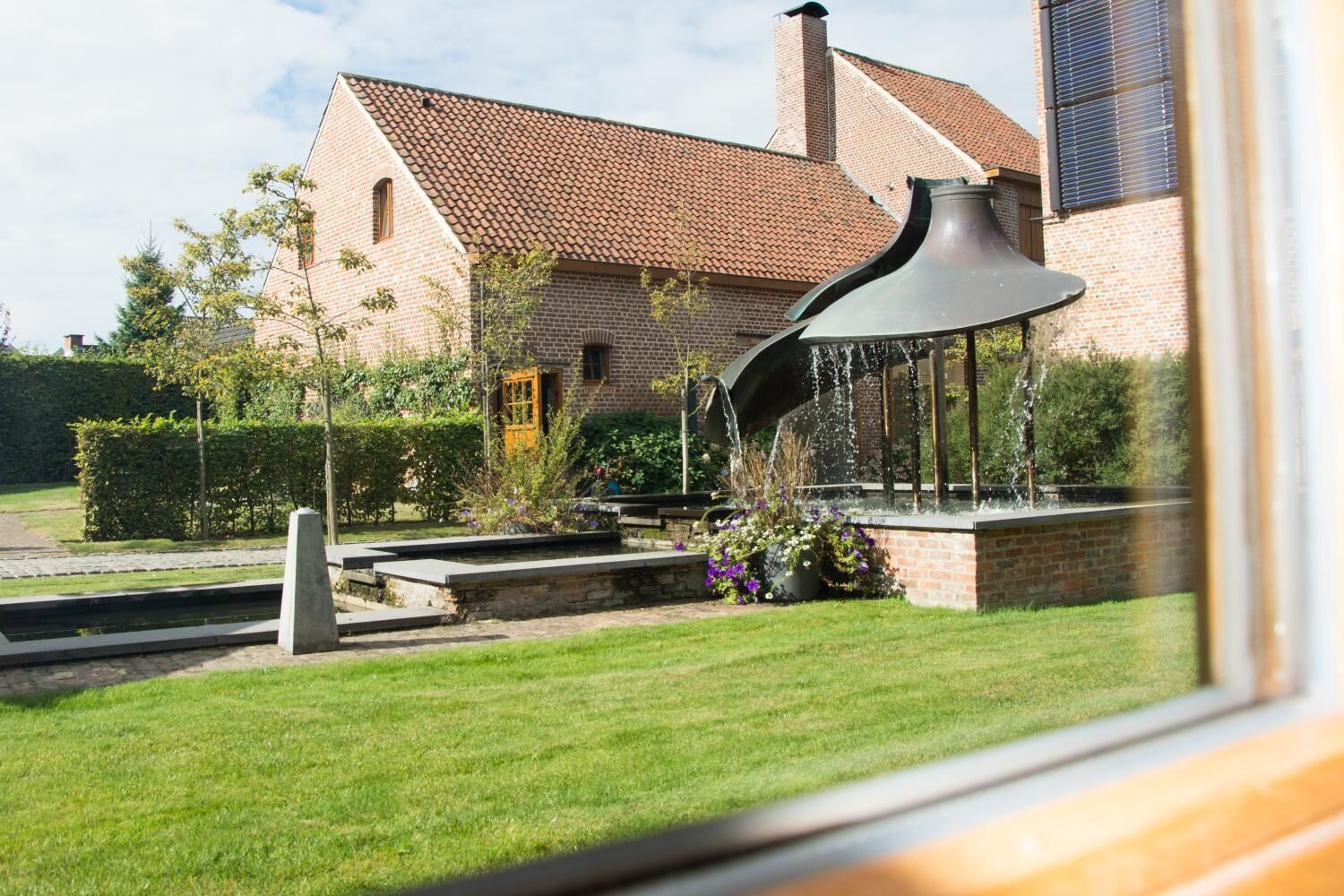 Una casa de ladrillo con techo de tejas, un jardín con fuente de agua y vista por la ventana.