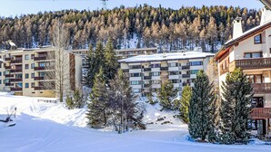A snow-covered residential area with multi-story buildings, including a hotel with balconies and a ski slope in the background.