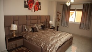 A bedroom with a large bed, two bedside tables, a nightstand, a decorative vase, and a window with curtains. - Lilia-appart (Oujda)
