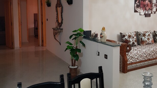 A spacious room with a large bed, a desk, and a chair. There are multiple shelves and a potted plant. - Lilia-appart (Oujda)