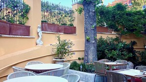 Un espace extérieur aménagé en terrasse, avec des tables rondes et des chaises en osier, entouré d’un petit muret et d’un grand arbre.