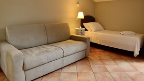 Une petite chambre d’hôtel composée d’une seule pièce, avec un canapé beige, un lit simple, une table de chevet avec une lampe et un mur sobre.