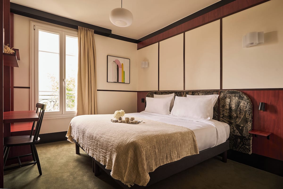A hotel room with a bed, a desk, a chair, a window with curtains, a wall-mounted artwork, and a bedside table.