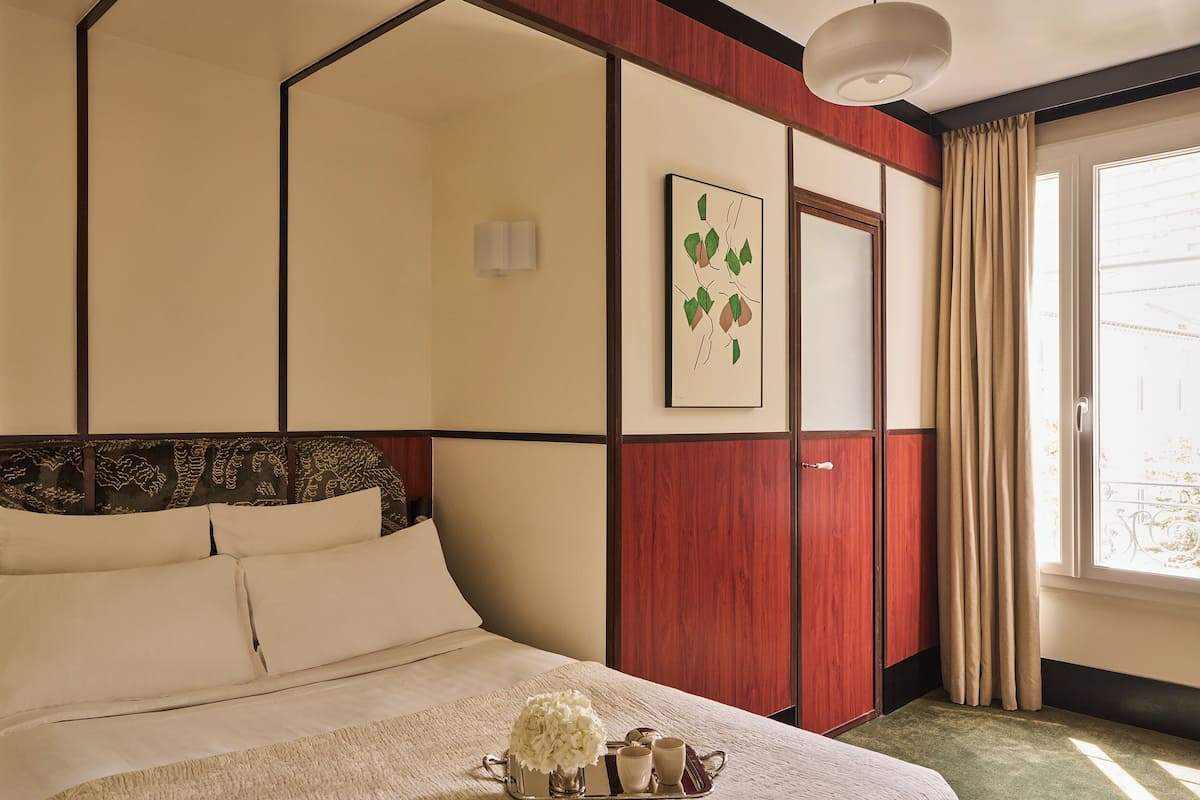 A hotel room with a bed, a bedside table with a tray of items, a window with curtains, and a wall-mounted picture.