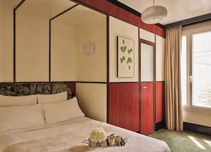 Quarto de hotel com uma cama, mesa de cabeceira com um porta-objetos, janela com cortinas e um quadro pendurado na parede.
