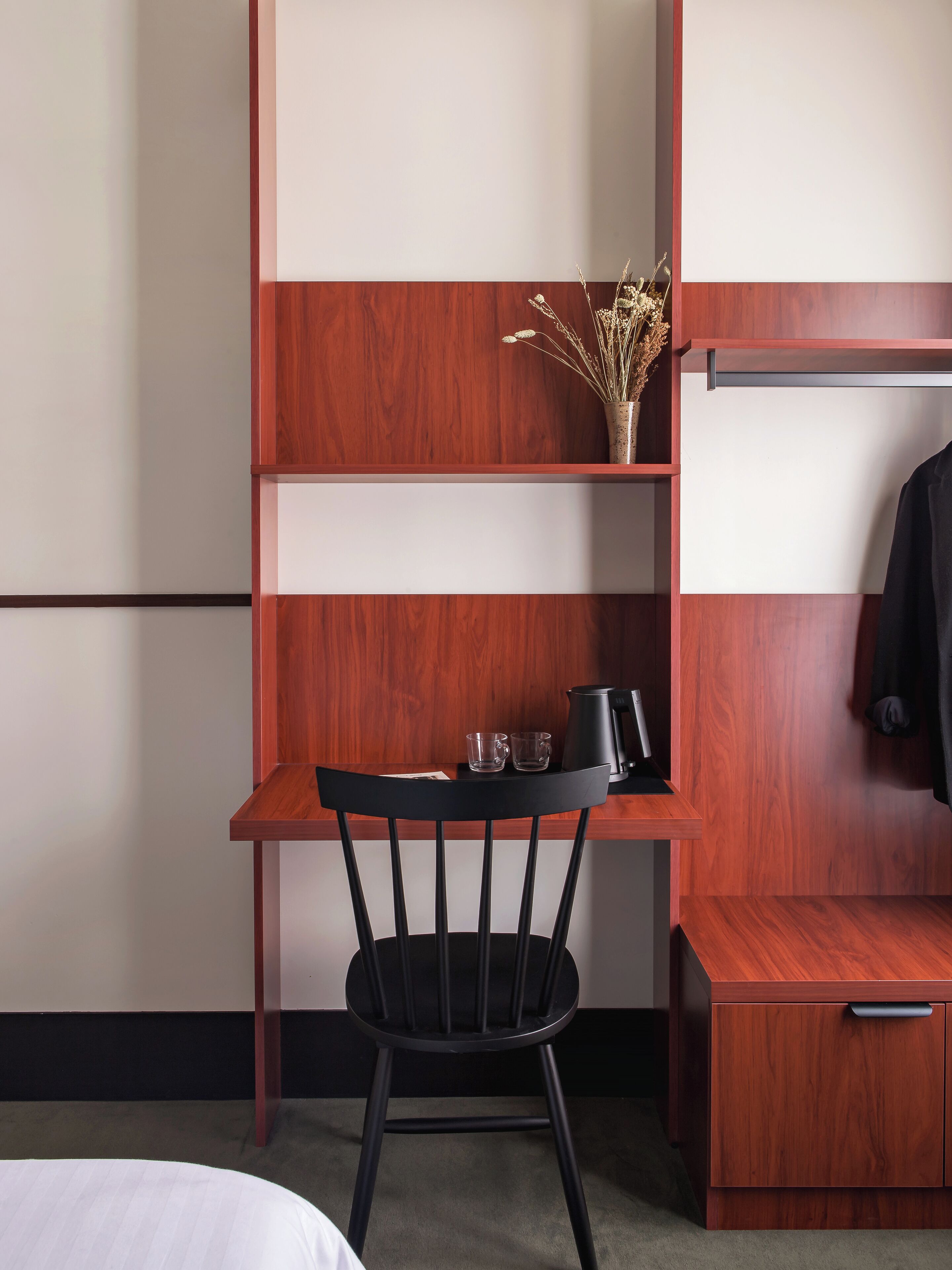 A compact hotel room with a wooden desk, a black chair, a small shelf with a vase, and a coat rack.