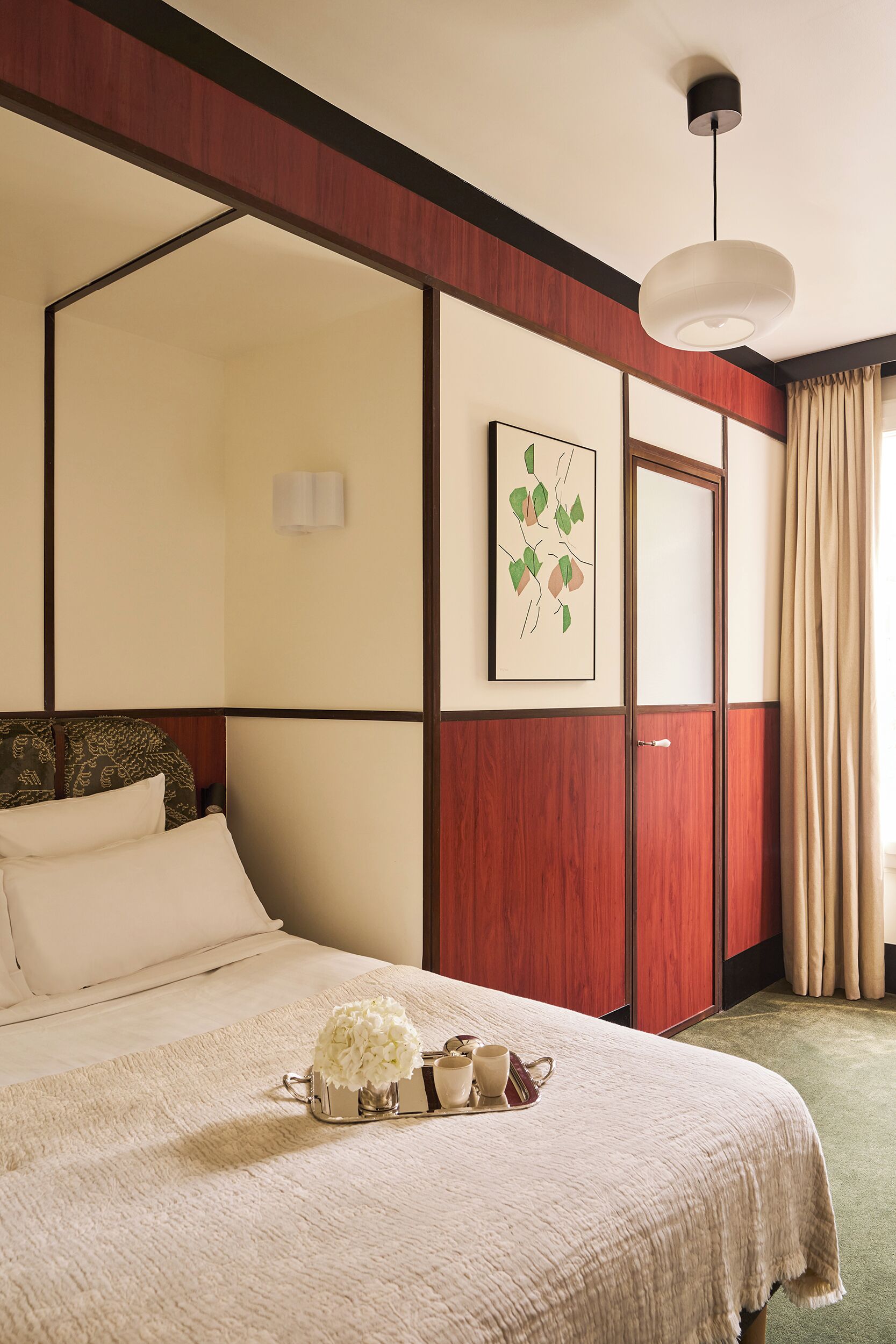 A hotel room with a bed, a bedside table with a tray of toiletries, a wall-mounted light fixture, and a framed artwork.