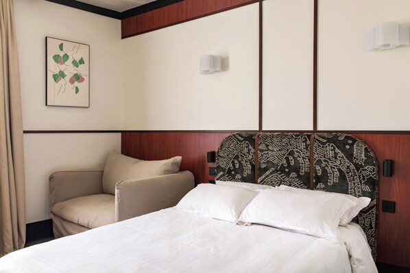 Una habitación de hotel con una cama, una silla, un cabecero con diseño estampado, una lámpara de pared y un cuadro enmarcado.