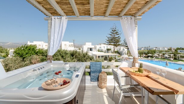 En takterrass med ett bubbelbad, ett träbord med frukt och glas, och utsikt över vita byggnader och en pool.