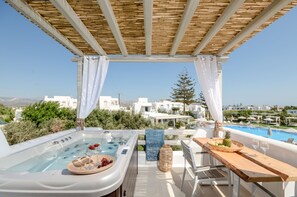 A rooftop terrace with a hot tub, a wooden table with fruits and glasses, and a view of white buildings and a pool. - Naxos Nature Suites (Naxos)
