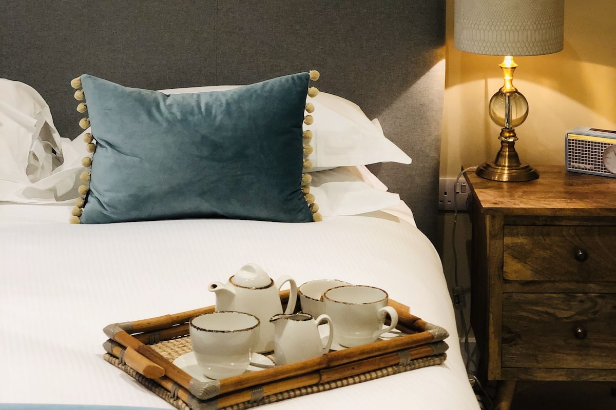 A neatly made bed with a tray of tea items, a lamp, and a painting on the wall.