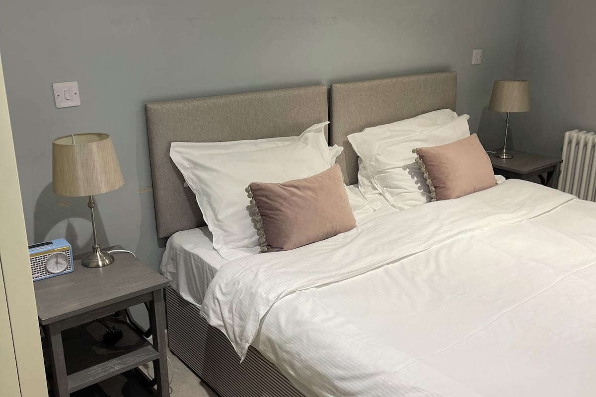 A neatly made bed with white and pink pillows, a bedside table with a lamp, and a radiator.