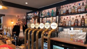 Um balcão de bar com diversas torneiras identificadas com tipos diferentes de bebidas alcoólicas, um balcão com copos e prateleiras cheias de garrafas.